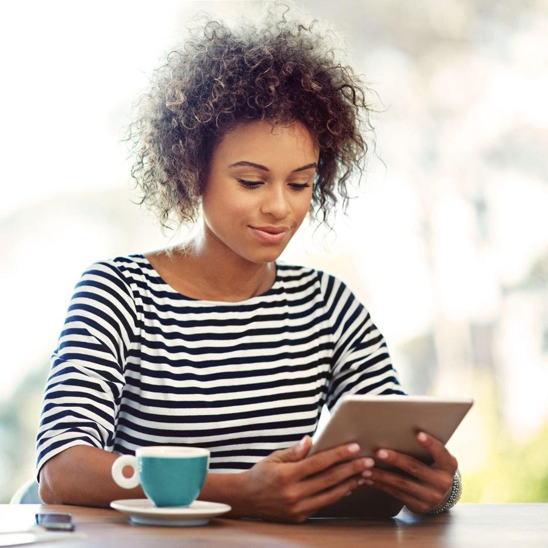 A smiling young woman with curly hair sitting at a table, enjoying a coffee while reading on her digital tablet.