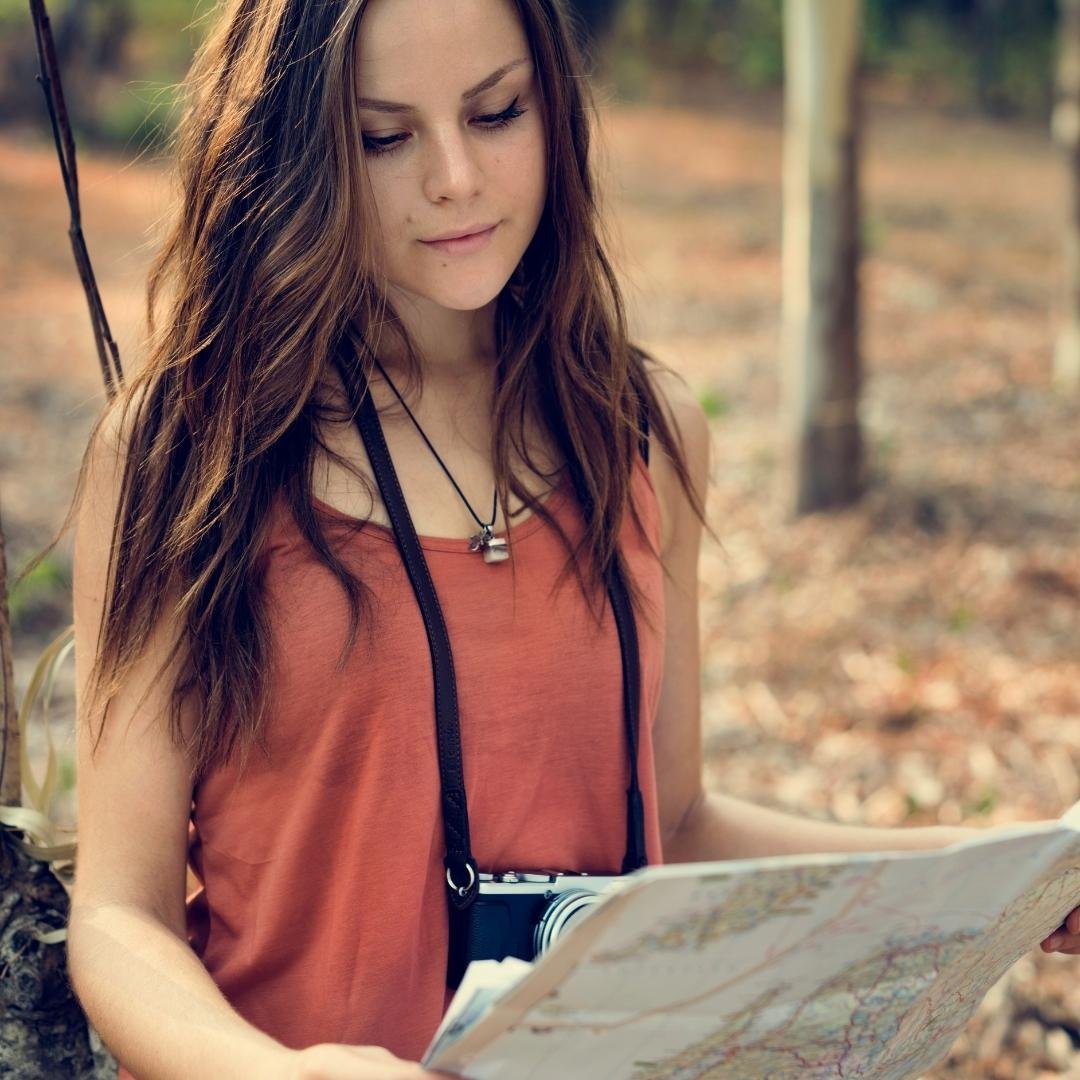 A young woman with long brown hair and a vintage camera around her neck, outdoors, looking down and focusing on an open paper map.