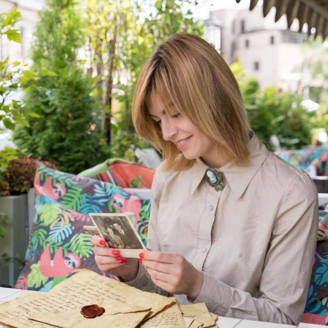 A blonde woman at an outdoor cafe looking fondly at a vintage black-and-white photograph, surrounded by old handwritten letters with wax seals.