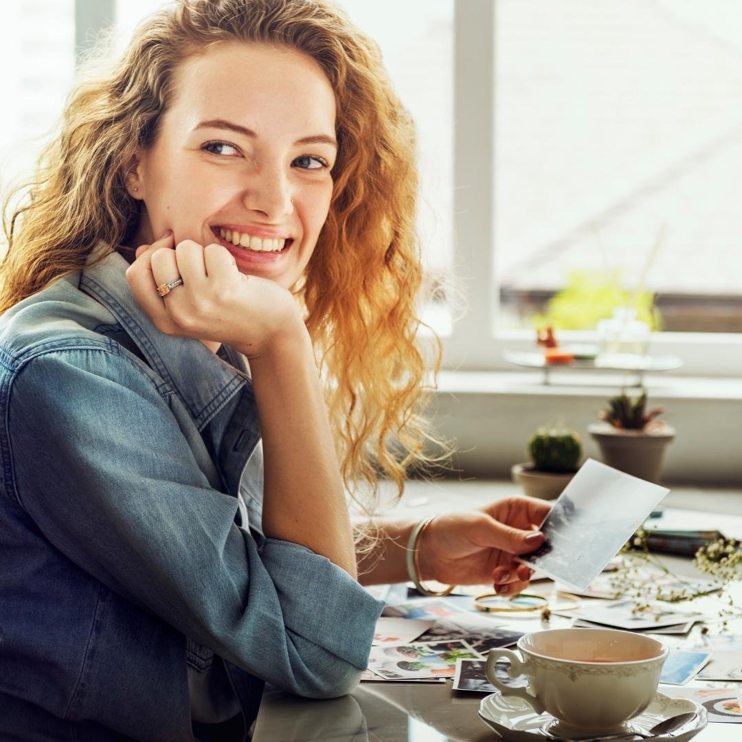 A cheerful curly-haired woman smiling by a bright window, holding a photo print with tea, plants, and memories scattered on the table.