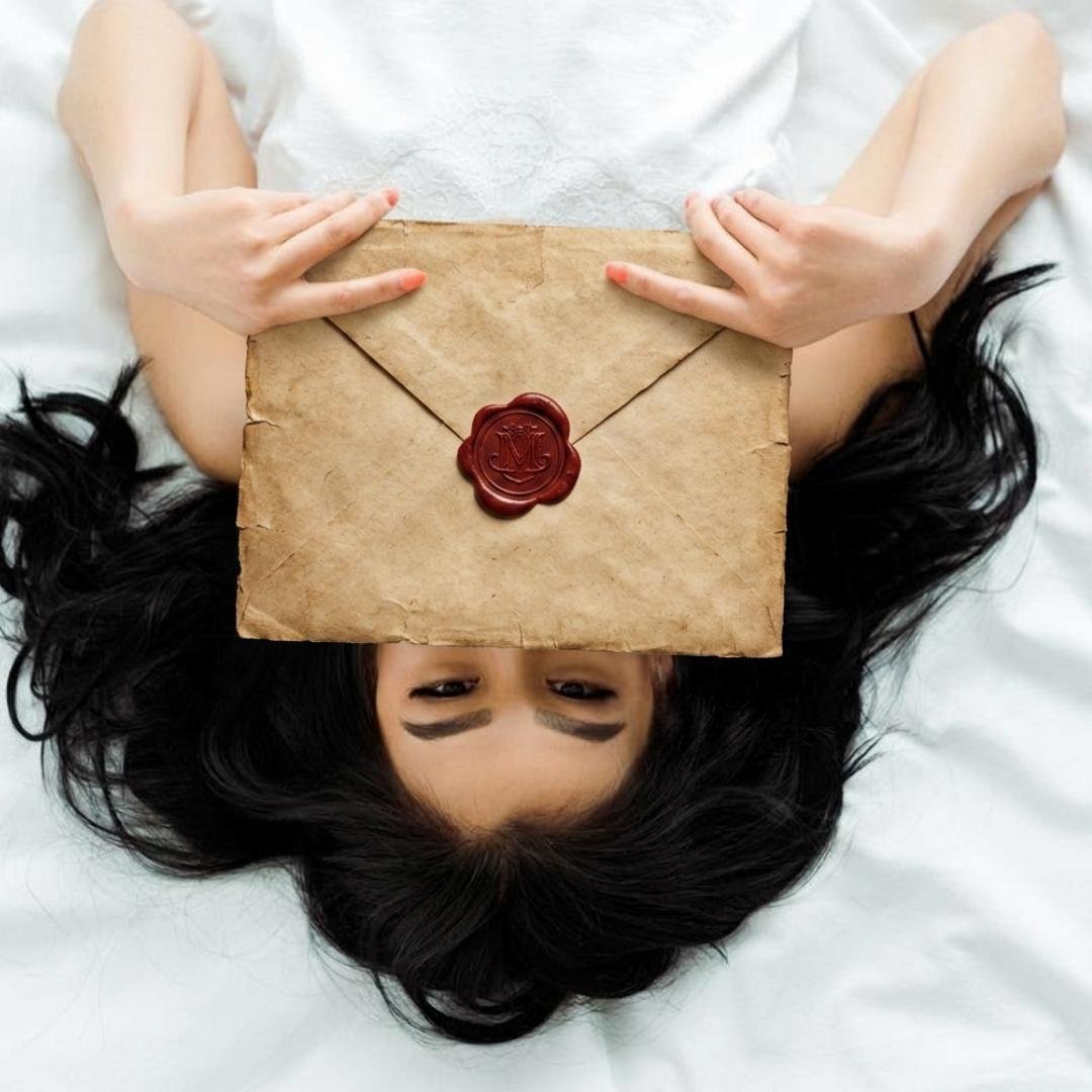 op-down view of a woman with long dark hair lying on white sheets, playfully holding a large vintage parchment envelope with a classic red wax seal over her face.